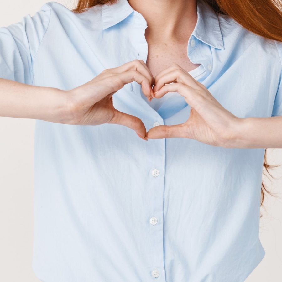 Young smiling redhead female showing heart sign and looking heartfelt at camera, standing over white background.